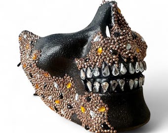 May include: A black face mask featuring a textured surface adorned with numerous small rose gold, amber, and clear crystals. The mask includes a prominent jawline design with clear, teardrop-shaped crystals arranged to resemble teeth along the mouth area.