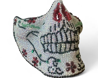 May include: A face mask covered in iridescent white rhinestones featuring a skull design. The skull is outlined in black rhinestones with red rhinestone floral accents and green rhinestone leaves. The teeth are defined by black rhinestones.