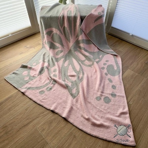 May include: A pink and gray butterfly-patterned blanket. The blanket features a large butterfly design in gray on a pink background. The blanket is folded over a surface, with the corner showing the logo "SiSous".