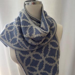 Könnte beinhalten: Blau-grauer Strickschal mit geometrischem Muster, drapiert auf einer Schaufensterpuppe. Der Schal hat ein sich wiederholendes blattartiges Design in Hellgrau auf blauem Hintergrund. Der Schal ist aus weichem Garn gefertigt und ein gemütliches Accessoire.