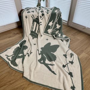 May include: A cream-coloured knitted blanket with a green fairy and lantern design. The blanket features a green border and decorative ivy and butterfly patterns. The blanket is draped over a wooden surface near a window.