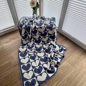 Könnte beinhalten: Eine gehäkelte Decke in Weiß und Marineblau mit einem Herzmuster. Die Decke ist gefaltet und liegt auf einem Holzboden.