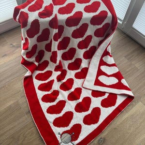 May include: A knitted blanket in red and white, patterned with red hearts on a white base. The blanket has a red border and is folded. The material appears soft and cosy, perfect for snuggling up with.