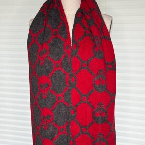 Puede incluir: Una bufanda tejida que presenta un patrón geométrico repetitivo de calaveras y formas de diamante interconectadas. La bufanda muestra un esquema de color reversible con un lado que muestra calaveras rojas sobre un fondo gris oscuro y el otro lado calaveras gris oscuro sobre un fondo rojo.