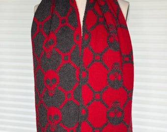 May include: A knitted scarf featuring a repeating geometric pattern of skulls and interconnected diamond shapes. The scarf displays a reversible color scheme with one side showing red skulls on a dark gray background and the other side showing dark gray skulls on a red background.