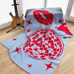Könnte beinhalten: Eine hellblaue Strickdecke mit einer großen roten Discokugel, einem roten Kussmund und roten vierzackigen Sternen. Die Decke hat einen roten Rand und enthält ein kleines Logo in der unteren Ecke mit dem Text SiSous pattern design.