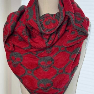 May include: A red and gray knitted scarf with a geometric pattern. The scarf is draped over a mannequin, showcasing its design and texture. The pattern features gray shapes against a red background, creating a visually striking accessory.