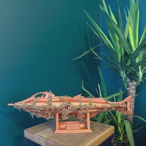 May include: A wooden model of the Nautilus submarine, with intricate details and a reddish-brown finish. The model is adorned with green moss and rests on a wooden stand. The word "NAUTILUS" is visible on the stand.