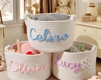 May include: Three round, off-white woven rope storage baskets with integrated handles. Each basket features a personalized name stitched in yarn: blue for Celine, pink for Olivia, and purple for Lucy, with a small flower accent on the Lucy basket.