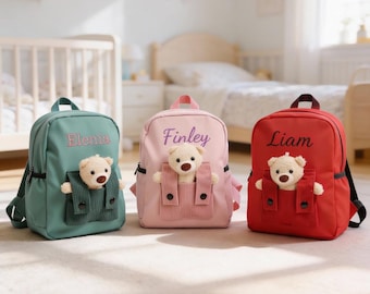 May include: Three personalized backpacks in teal, pink, and red, each with a teddy bear in the front pocket. The teal backpack has "Elenia" embroidered, the pink one says "Finley", and the red one says "Liam".