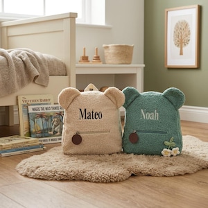 Pode incluir: Duas mochilas de pelúcia em forma de urso feitas de tecido fleece texturizado. A mochila bege apresenta o nome bordado Mateo, enquanto a mochila verde-azulado apresenta o nome bordado Noah e um pequeno detalhe de flor em crochê na frente.