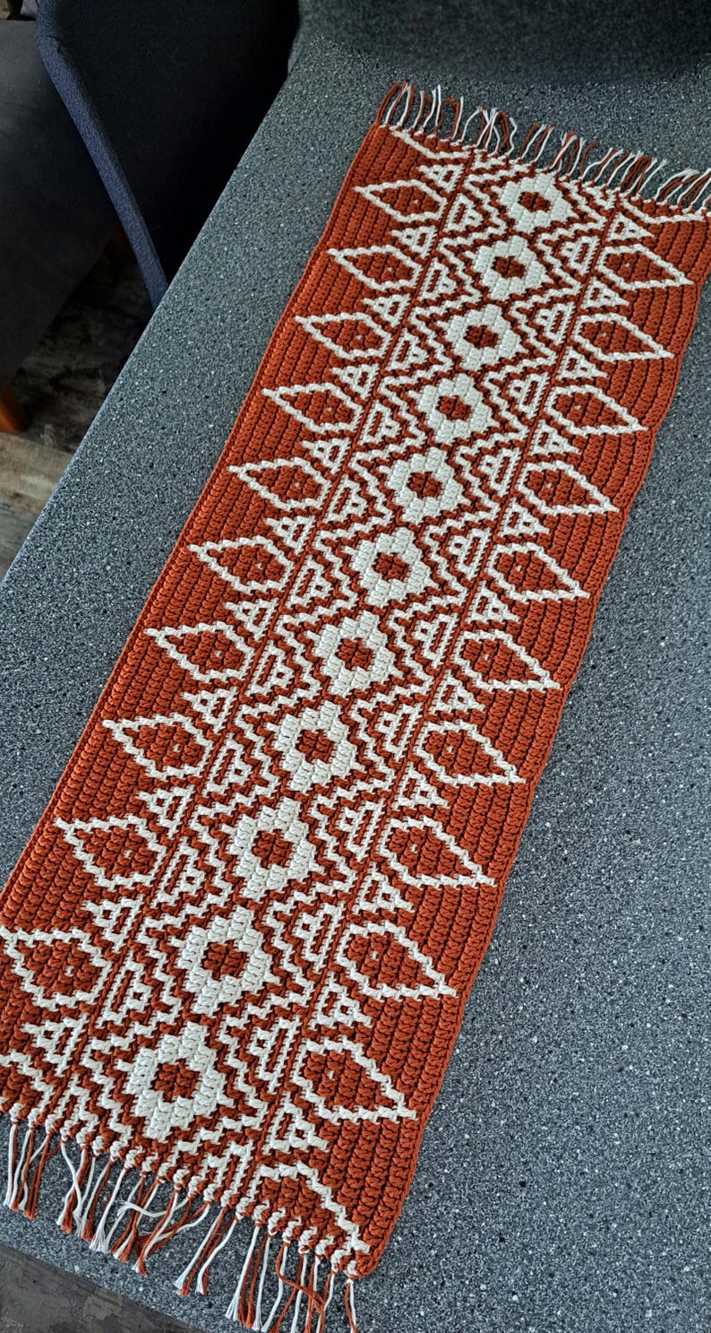 A rectangular, hand-crocheted table runner in a burnt orange color with a repeating geometric pattern in white. The runner has a fringed edge and is made of cotton or a similar yarn. The design features diamond and floral shapes.
