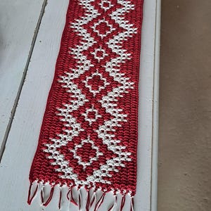 Puede incluir: Camino de mesa rectangular, tejido a mano, en rojo y blanco. El camino de mesa presenta un patrón geométrico repetitivo y flecos en ambos extremos. El camino de mesa mide aproximadamente 30 cm de ancho y 76 cm de largo.