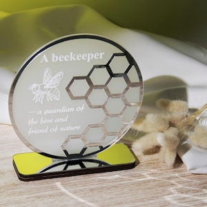 Könnte beinhalten: Dekorative, runde Plakette mit dem Text "A beekeeper - a guardian of the hive and friend of nature". Sie zeigt eine Bienenillustration und ein Wabenmuster. Sie steht auf einem rechteckigen Sockel mit gelbem Akzent.