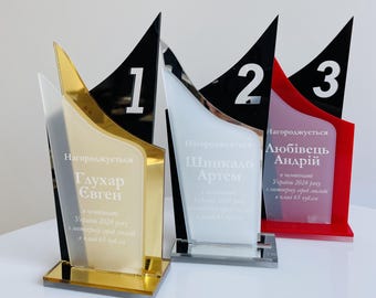 Könnte beinhalten: Vier Acryl-Trophäen mit modernem Design. Die Trophäen haben einen goldenen, silbernen und roten Sockel mit einer schwarzen dreieckigen Spitze. Jede Trophäe hat eine Nummer von 1 bis 3 und Text in einer Fremdsprache.