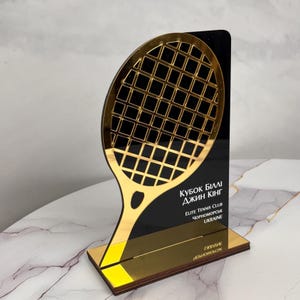 Puede incluir: Un trofeo de tenis dorado y negro. El trofeo presenta un diseño de raqueta de tenis dorada sobre un fondo negro. El texto en el trofeo dice "KUBOK BILLI DJIN KING" y "ELITE TENNIS CLUB CHORNOMORSK UKRAINE".
