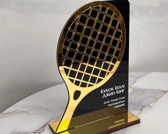 May include: A gold and black tennis-themed trophy. The trophy features a gold tennis racket design against a black background. Text on the trophy reads "KUBOK BILLI DJIN KING" and "ELITE TENNIS CLUB CHORNOMORSK UKRAINE".
