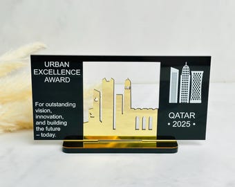 Puede incluir: Un premio Urban Excellence negro y dorado. El premio presenta una silueta dorada de paisaje urbano sobre un fondo transparente, con el texto "URBAN EXCELLENCE AWARD" y "QATAR • 2025." El premio está sobre una base negra.