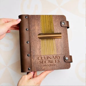 Puede incluir: Un libro de recetas de madera con encuadernación de cuero marrón y las palabras "CULINARY SECRETS Madison" grabadas en la portada. El libro contiene varias formas de pasta y un dispositivo Dyson de color cobre.