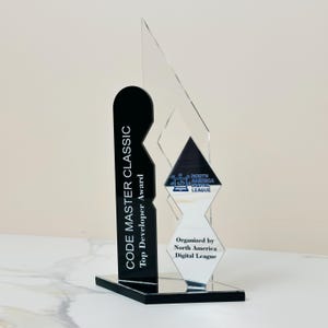 Op de afbeelding: Een heldere acryl trofee met een zwarte basis en accenten. De trofee bevat de tekst "CODE MASTER CLASSIC Top Developer Award" en "Organized by North America Digital League."