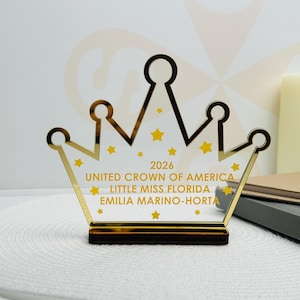 May include: A gold-coloured crown-shaped award with the text "2026 UNITED CROWN OF AMERICA LITTLE MISS FLORIDA EMILIA MARINO-HORTA" and yellow star accents. The crown sits on a dark brown base.