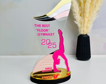 May include: A pink and gold acrylic award with a silhouette of a gymnast performing a backbend. The award reads "The Best "Floor" Gymnast 2025" and "Local gymnastics competitions North Carolina".