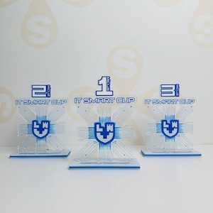 Puede incluir: Tres trofeos de acrílico transparente con detalles azules. Cada trofeo presenta un diseño de placa de circuito, un emblema de escudo y el texto "IT SMART CUP". Los trofeos están numerados 1, 2 y 3, indicando la clasificación.