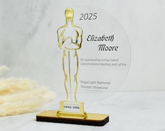 Könnte beinhalten: Eine goldfarbene Acryl-Auszeichnung in Form einer Statue, montiert auf einem rechteckigen Sockel. Die Auszeichnung trägt die Aufschrift "2025", "Elizabeth Moore" und "StageLight National Theater Showcase". Der Sockel ist dunkelbraun.