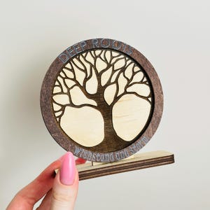 May include: A wooden tree of life decoration with the words "DEEP ROOTS" and "WITHSTAND THE STORM" etched into the design. The tree is light-colored wood, set within a dark brown circular frame and base.