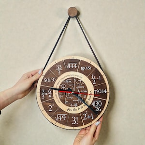 Op de afbeelding: Een ronde houten klok met een donkerbruine wijzerplaat met wiskundige vergelijkingen en getallen. De klok heeft zwarte wijzers met een rode secondewijzer en wordt opgehangen door een zwarte band. De tekst "For the best math teacher" is zichtbaar.