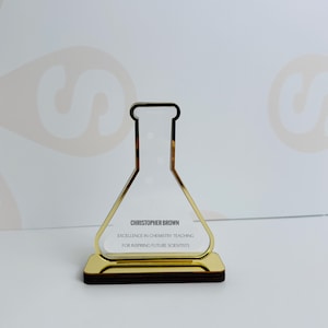Può includere: Un premio in acrilico trasparente con bordi dorati a forma di beuta da chimica. Il premio poggia su una base marrone scuro. Il testo sul premio recita "CHRISTOPHER BROWN, EXCELLENCE IN CHEMISTRY TEACHING, FOR INSPIRING FUTURE SCIENTISTS."