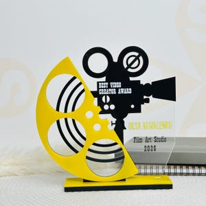 Puede incluir: Un trofeo del premio al Mejor Creador de Vídeos. El premio presenta una bobina de película amarilla y una silueta de cámara de cine negra. El texto del premio dice "BEST VIDEO CREATOR AWARD", "OLYA KOVALENKO", "Film Art Studio" y "2026".