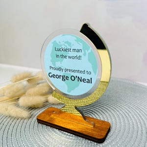 Puede incluir: Un premio decorativo en forma de globo con una base de madera. El globo presenta un mapa del mundo azul claro y el texto "Luckiest man in the world! Proudly presented to George O'Neal".