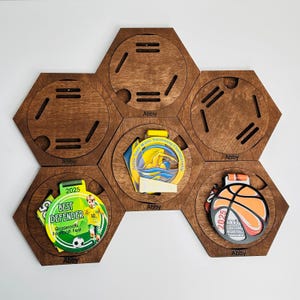 May include: A wooden medal display in a hexagonal design, featuring six compartments. It showcases medals from swimming, basketball, and football. The medals are inscribed with the name "Abby" and the year 2025. The football medal says "Best Defender".