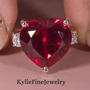 May include: A gold-toned ring featuring a large, heart-shaped, faceted red gemstone. The ring has smaller, clear gemstones on either side of the heart. The text "KylieFineJewelry" is at the bottom.