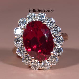 May include: A rose gold ring with a large, oval-cut, deep red gemstone encircled by a halo of smaller, clear gemstones. The ring is set against a soft pink background, with the text "KylieFineJewelry" visible.