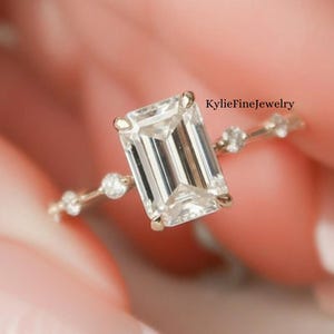 Könnte beinhalten: Ein goldener Ring mit einem Smaragd-Schliff-Diamanten als Mittelstück, flankiert von kleineren, runden Diamanten entlang des Bands. Der Ring wird vor einem weichen Hintergrund präsentiert, um sein Funkeln und Design hervorzuheben.