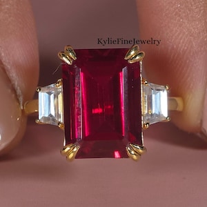 May include: A gold ring with a large, rectangular, deep red gemstone, and two smaller, rectangular, clear gemstones. The ring is set against a pink background. The text "KylieFineJewelry" is visible.