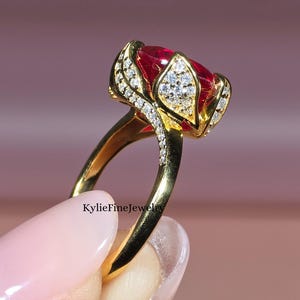 May include: A gold ring featuring a large, oval-cut red gemstone, accented with smaller white gemstones. The ring's setting has a floral design. The ring is held up against a soft pink background. The text "KylieFineJewels" is visible.