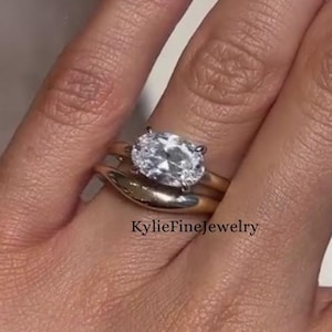 May include: A gold ring set with a large oval-cut diamond, paired with a simple gold band. The rings are worn on a finger with long, polished nails. The text "KylieFineJewelry" is visible.
