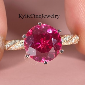 May include: A gold ring featuring a large, round, pink gemstone with multiple facets. The band is decorated with small, clear stones. The ring is held between fingers against a pink backdrop. The text "KylieFineJewelry" is visible.