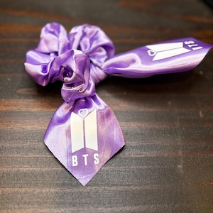 May include: A purple satin scrunchie with a white BTS logo printed on a ribbon. The scrunchie is knotted and has a decorative ribbon with the BTS logo and a heart symbol.
