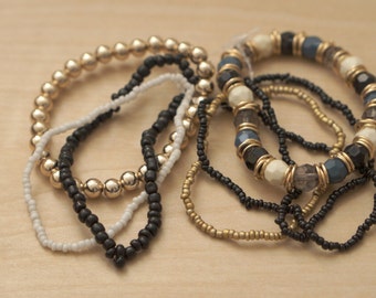 May include: A set of layered necklaces featuring gold, black, and white beads. The necklaces are made of different materials, including metal, glass, and plastic.