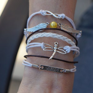 May include: A stack of bracelets on a person's wrist. The bracelets are white, brown, and black. The bracelets have charms, including a dragonfly, wings, and a charm with the word "faith" on it.