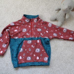 Könnte beinhalten: Ein rotes und tealfarbenes Fleece-Pullover mit einem Bären- und Pilzmuster. Das Oberteil hat eine tealfarbene Tasche und einen Kragen, einen roten Reißverschluss und ein Bären- und Pilzmuster. Das Kleidungsstück ist für Kinder.