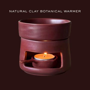 May include: A dark red clay botanical warmer with the text NATURAL CLAY BOTANICAL WARMER printed above. The cylindrical ceramic vessel features an open base containing a small, lit tea light candle inside a matching handled clay dish.