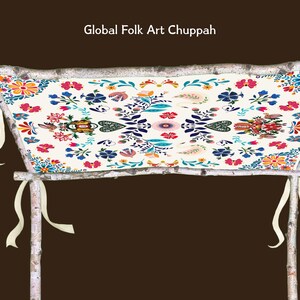 May include: A Global Folk Art Chuppah with a cream-colored fabric canopy adorned with colorful floral patterns in red, blue, and orange. The canopy is supported by birch poles and tied with cream-colored ribbons.