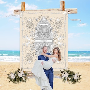 May include: A couple on a beach, with the groom carrying the bride. Behind them is a beige backdrop with a black and white illustration. The backdrop is supported by wooden beams. Floral arrangements are at the base of the backdrop.
