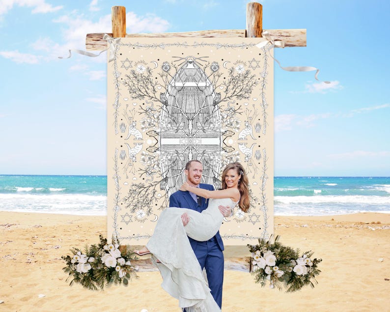 May include: A couple on a beach, with the groom carrying the bride. Behind them is a beige backdrop with a black and white illustration. The backdrop is supported by wooden beams. Floral arrangements are at the base of the backdrop.