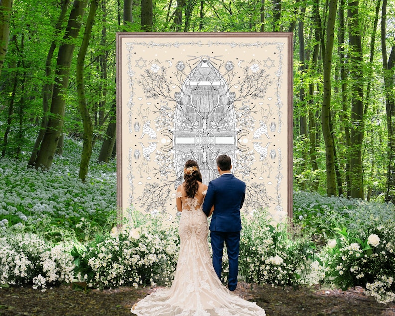 May include: A couple stands before a large framed artwork in a forest setting. The artwork features intricate black and white illustrations on a cream background. The bride wears a lace wedding dress, and the groom is in a blue suit. White flowers line the path.